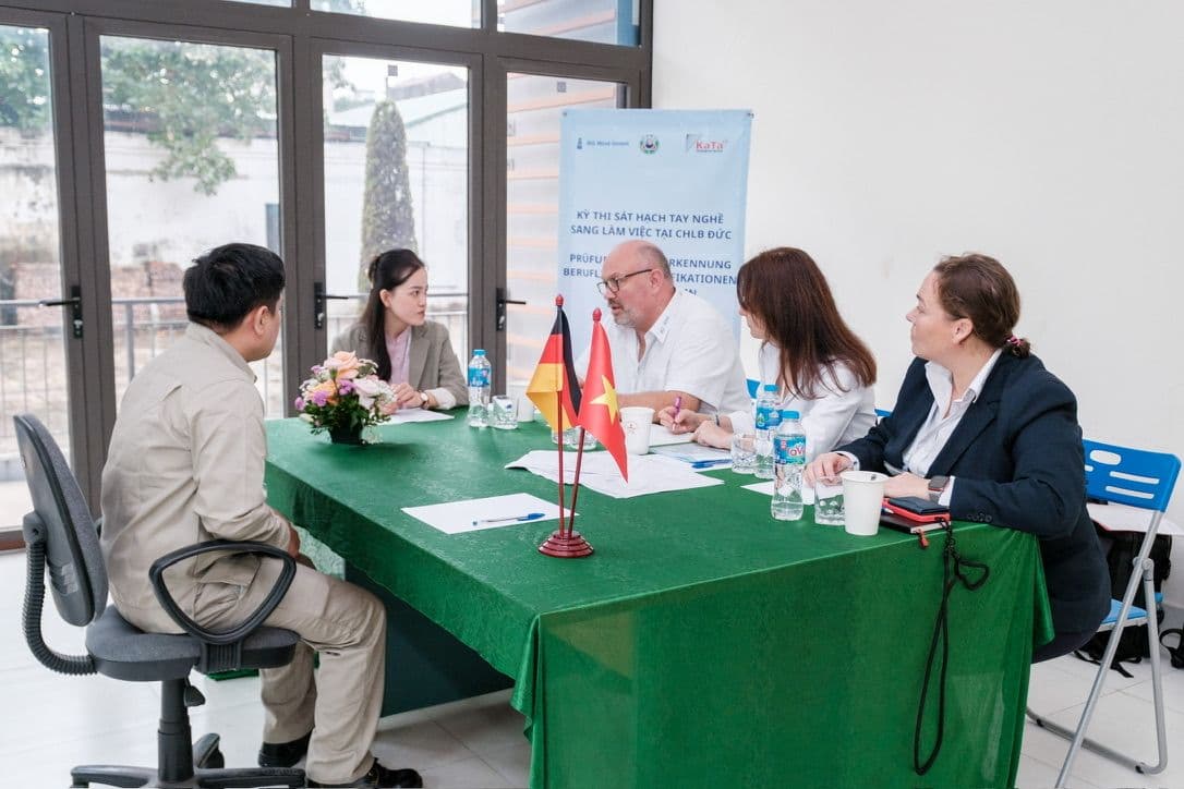 Auswahlgespräch: Kandidat, Prüfer aus Deutschland und Dolmetscherin an einem Tisch mit deutscher und vietnamesischer Flagge